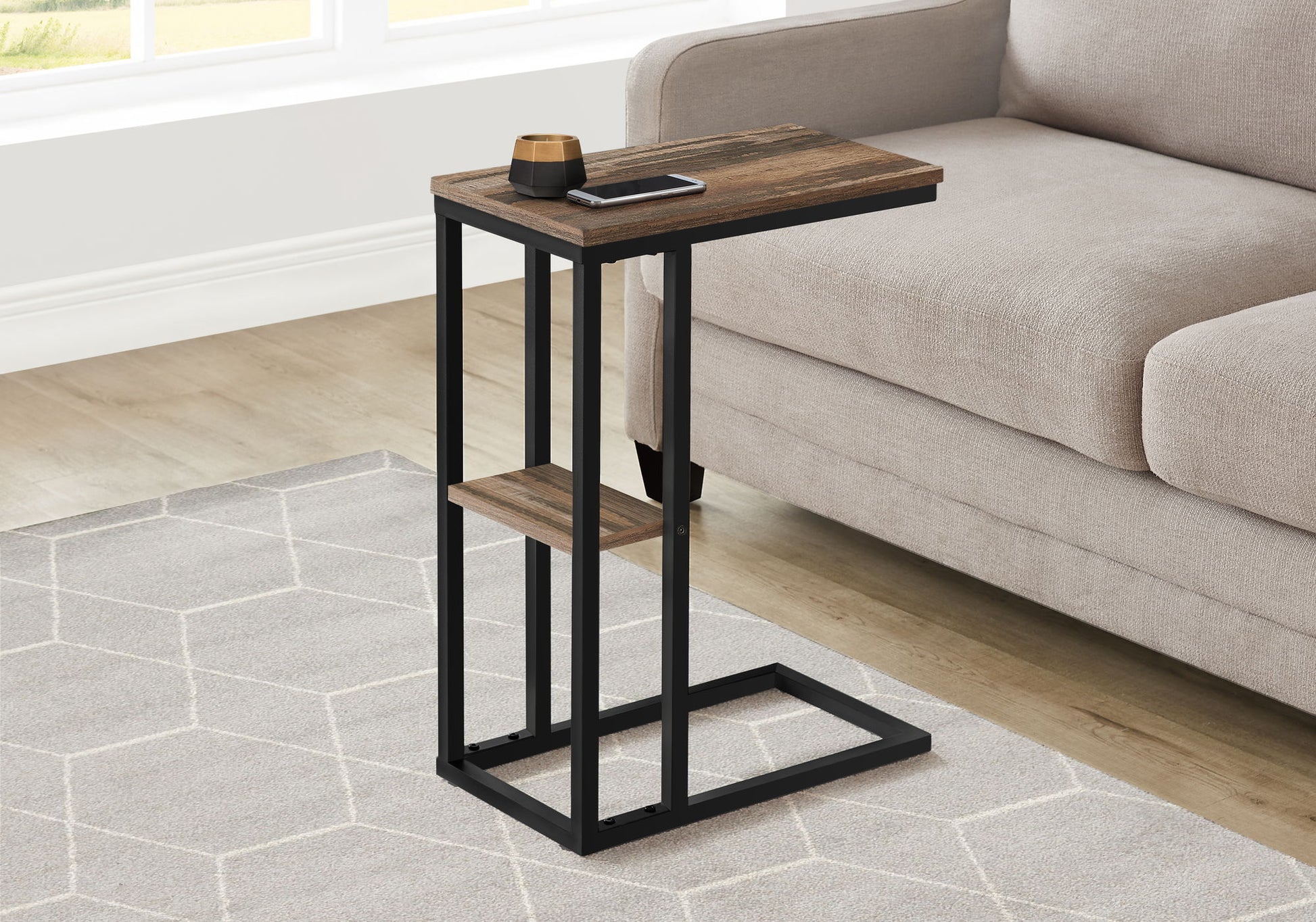 The Accent Table C-Shaped, Contemporary & Modern with a dark wood top and black metal frame sits next to a beige sofa on a light geometric-patterned rug, holding a phone and small cup.