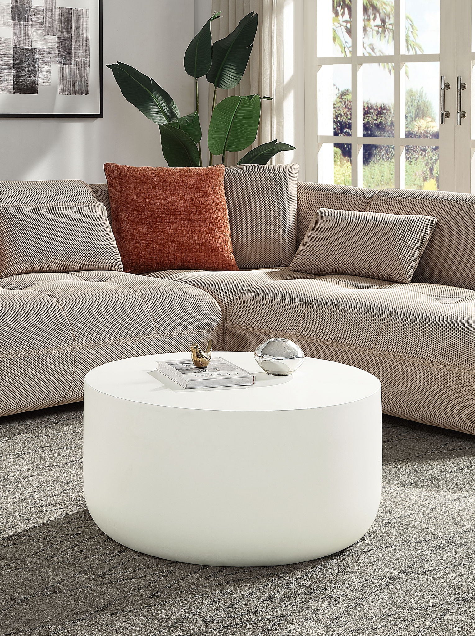 A modern living room with a light beige sectional sofa, rust-colored pillow, leafy plant, large windows, and the Dustan - Natural Coffee Table in white displaying decor items.