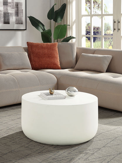 A modern living room with a light beige sectional sofa, rust-colored pillow, leafy plant, large windows, and the Dustan - Natural Coffee Table in white displaying decor items.