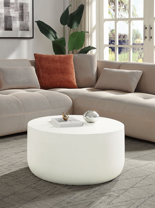 A modern living room with a light beige sectional sofa, rust-colored pillow, leafy plant, large windows, and the Dustan - Natural Coffee Table in white displaying decor items.