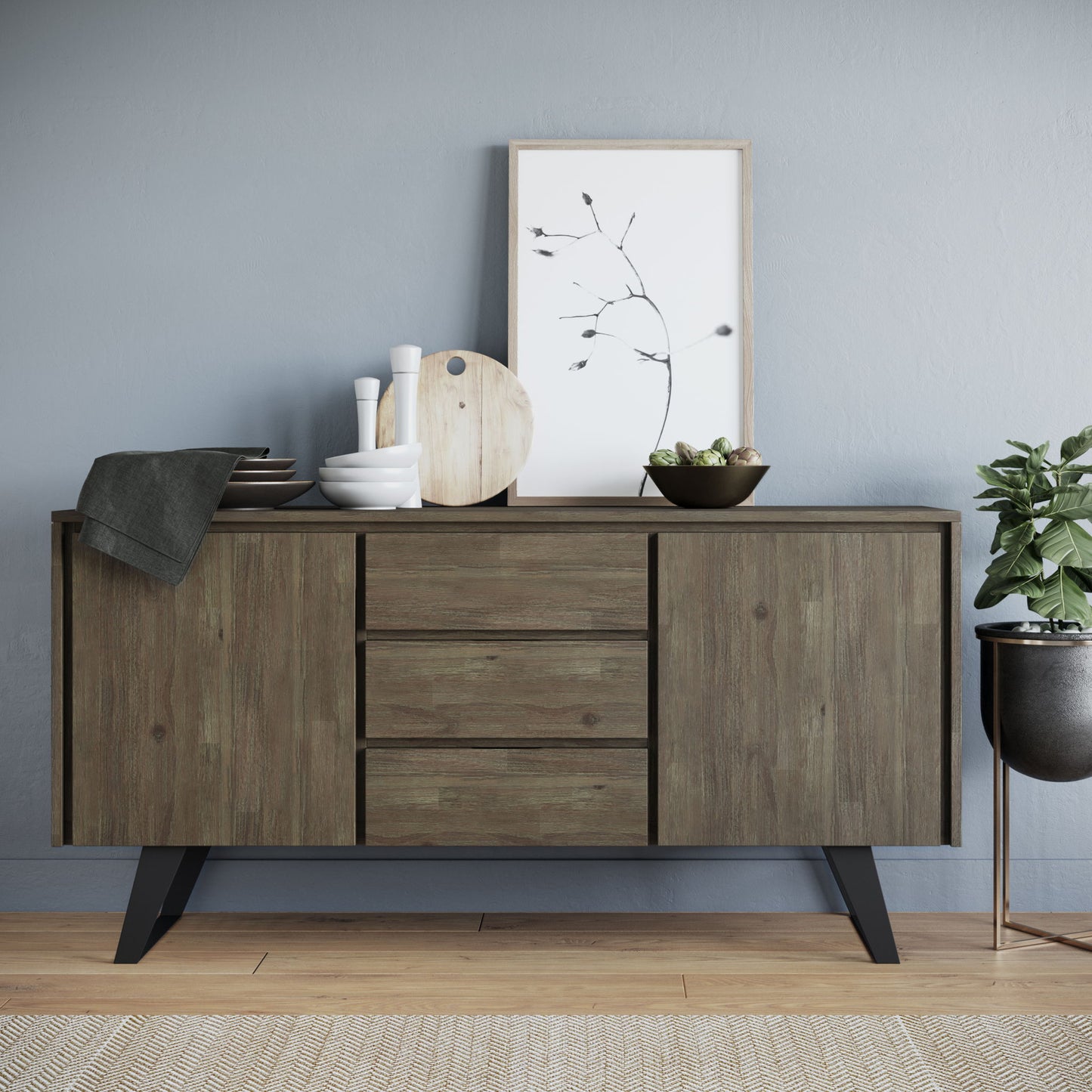 The Lowry - Handcrafted Sideboard Buffet, featuring three drawers and cabinets, stores dishes, bowls, a cutting board, and a framed botanical print. A potted plant sits on the floor to the right, while a textured rug covers the wooden flooring.