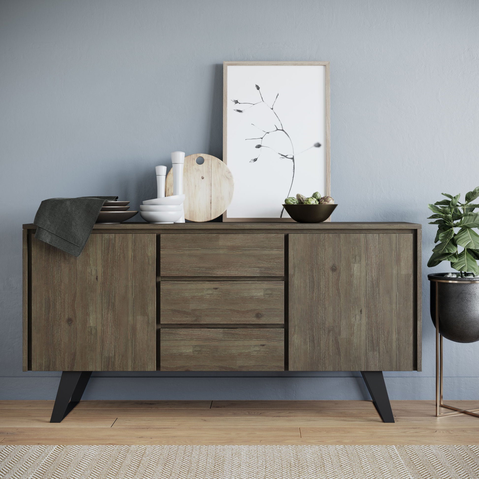 The Lowry - Handcrafted Sideboard Buffet, featuring three drawers and cabinets, stores dishes, bowls, a cutting board, and a framed botanical print. A potted plant sits on the floor to the right, while a textured rug covers the wooden flooring.
