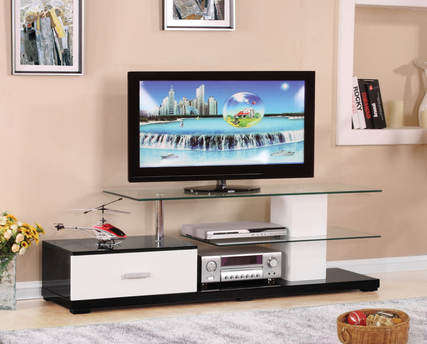The Ivana TV Stand in White/Black features a flat-screen TV, glass shelves for electronics, a drawer, a toy helicopter on the left, a basket of toys on the floor, and framed art on the beige wall.