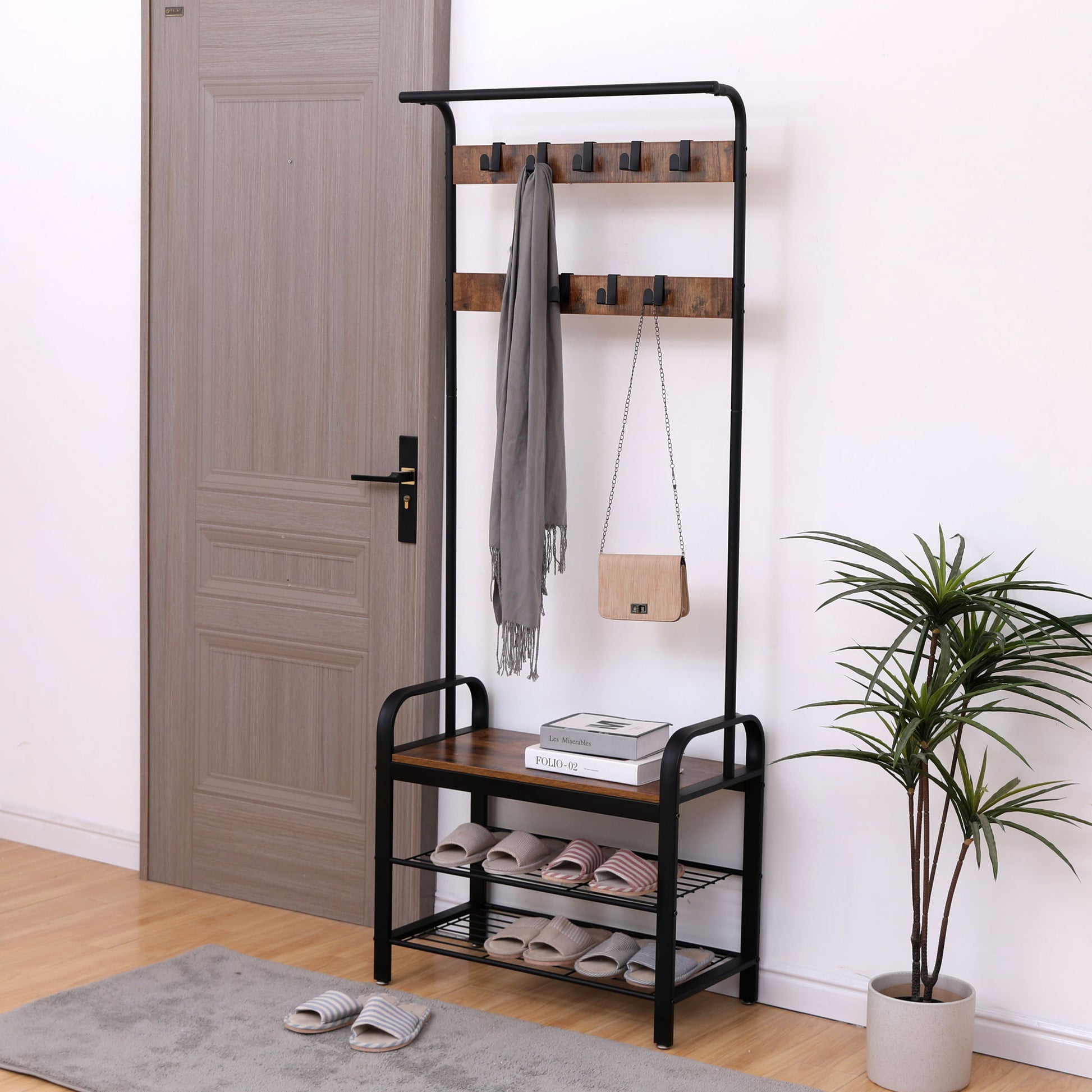 A Hall Tree With Shoe Bench For Entryway with 9 removable hooks holds a gray scarf and small bag, features shelves with slippers, a bench, books, a potted plant, and has a closed wooden door in the background.