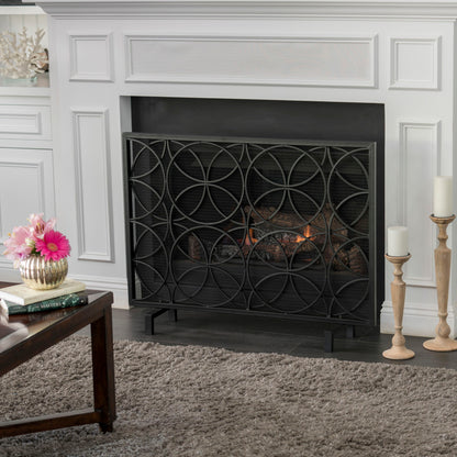 A modern living room features a white fireplace with a Thayer - Single Panel Iron Fireplace Screen With Geometric Design, a lit fire, gray rug, coffee table with flower vase, and two tall candle holders on the right.