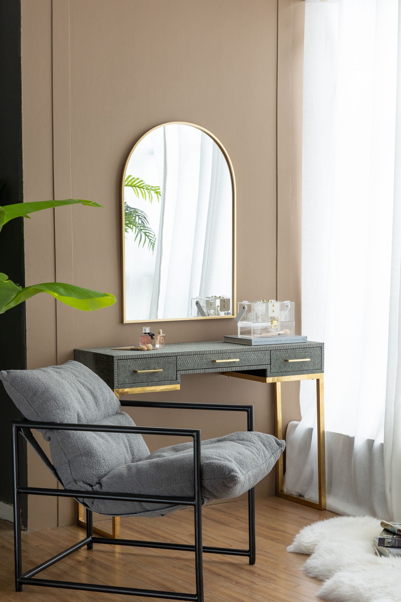 A modern vanity table with gold legs stands by a window with sheer curtains. An Arched Mirror With Metal Frame is above the table, paired with a gray cushioned chair and decorative items on the tabletop.