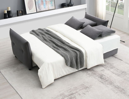 A modern living room features the Irina Pull Out Sleep With Storage, unfolded as a bed with white bedding, gray pillows, and a textured gray throw blanket. Art and decor sit on a shelf behind, sunlight streaming through large windows.