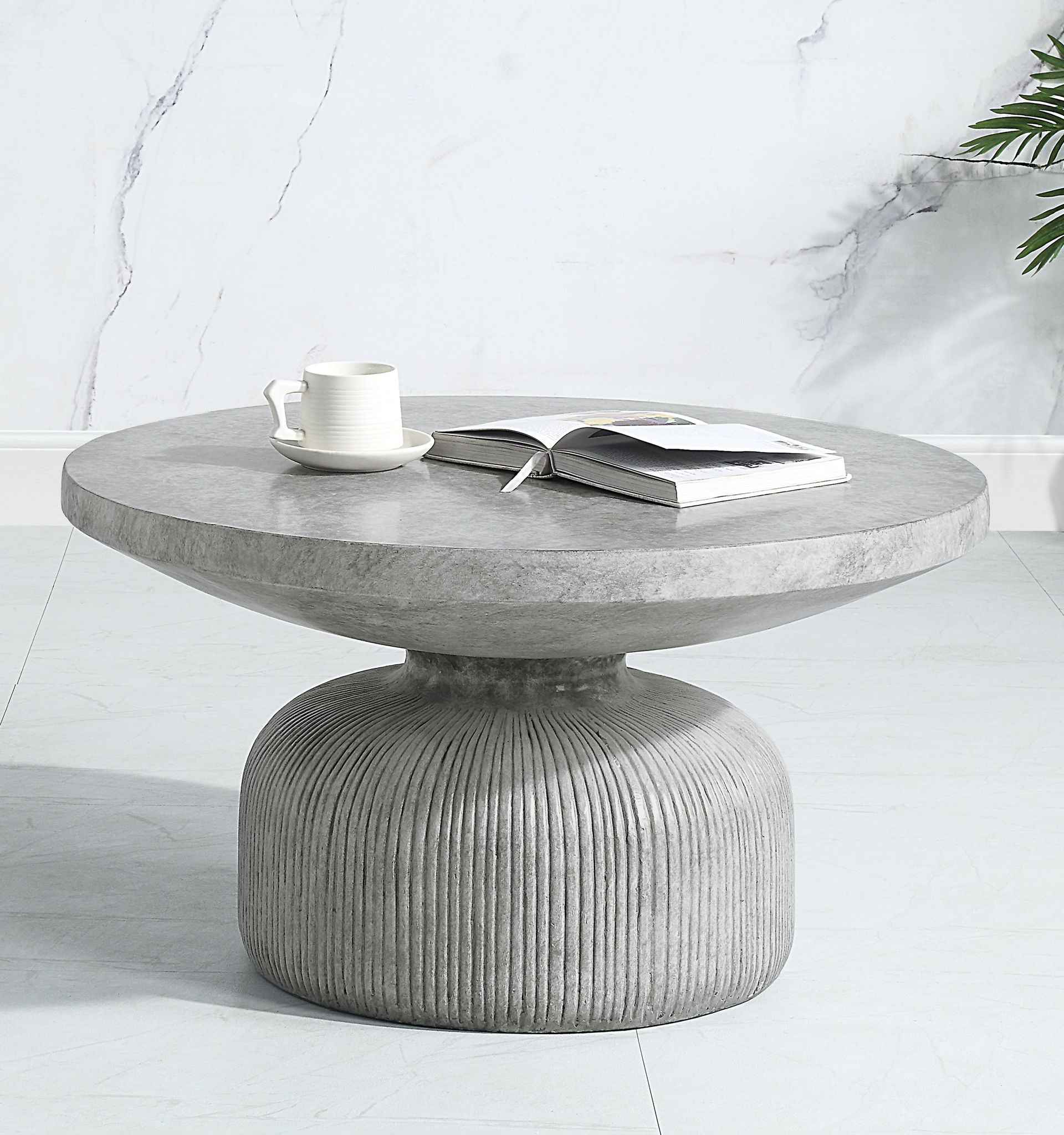 The Laddie - Weathered Table, a round modern gray concrete coffee table with a ribbed base, sits on a light floor. On top are an open book and white cup with saucer. A marble-patterned wall and green plant are in the background.