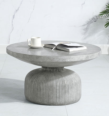 The Laddie - Weathered Table, a round modern gray concrete coffee table with a ribbed base, sits on a light floor. On top are an open book and white cup with saucer. A marble-patterned wall and green plant are in the background.