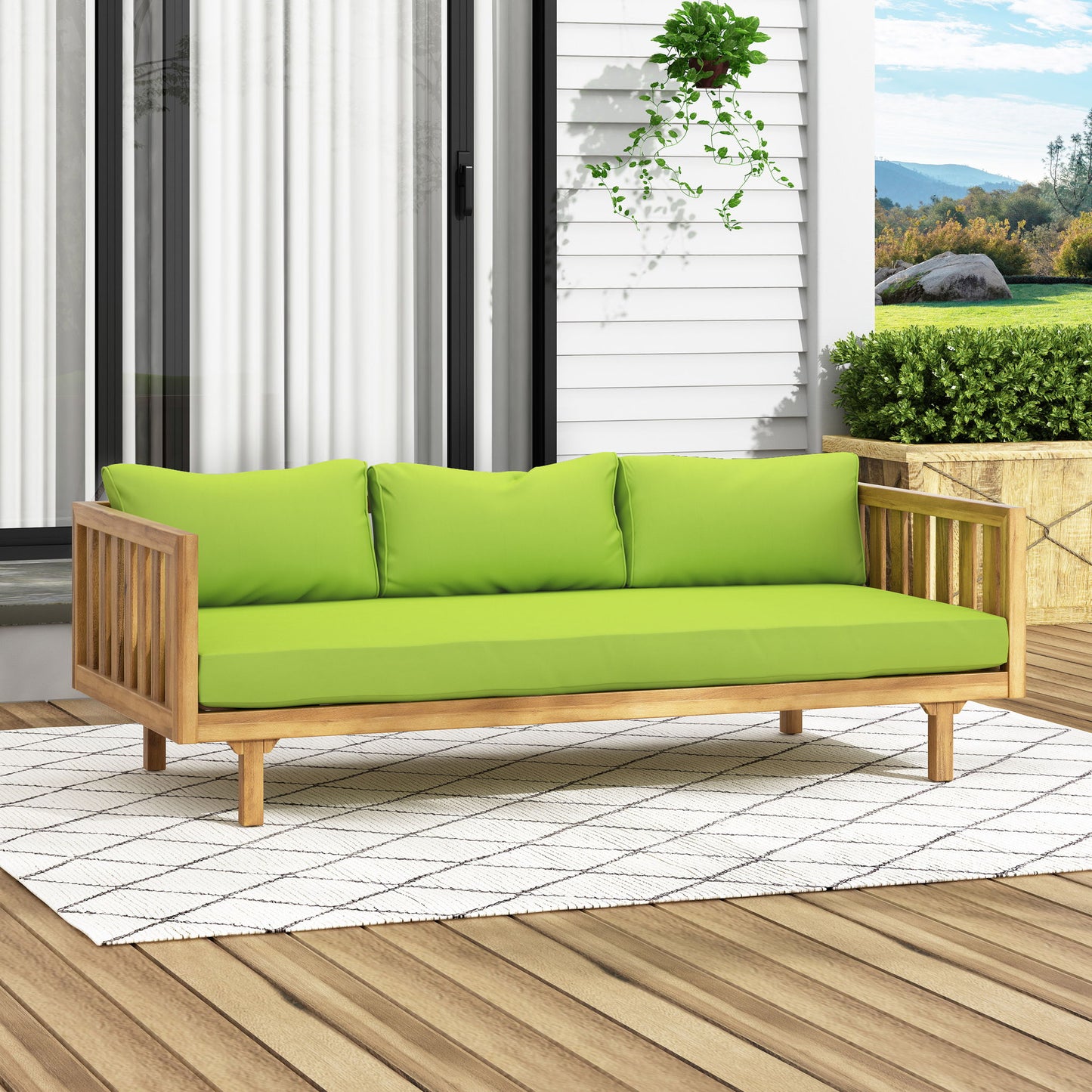 The Claremont - 3 Seater Daybed, crafted from teak and acacia wood with water-resistant green cushions and three pillows, rests on a white patterned rug on a wooden deck surrounded by plants, with a house and mountains in the background.