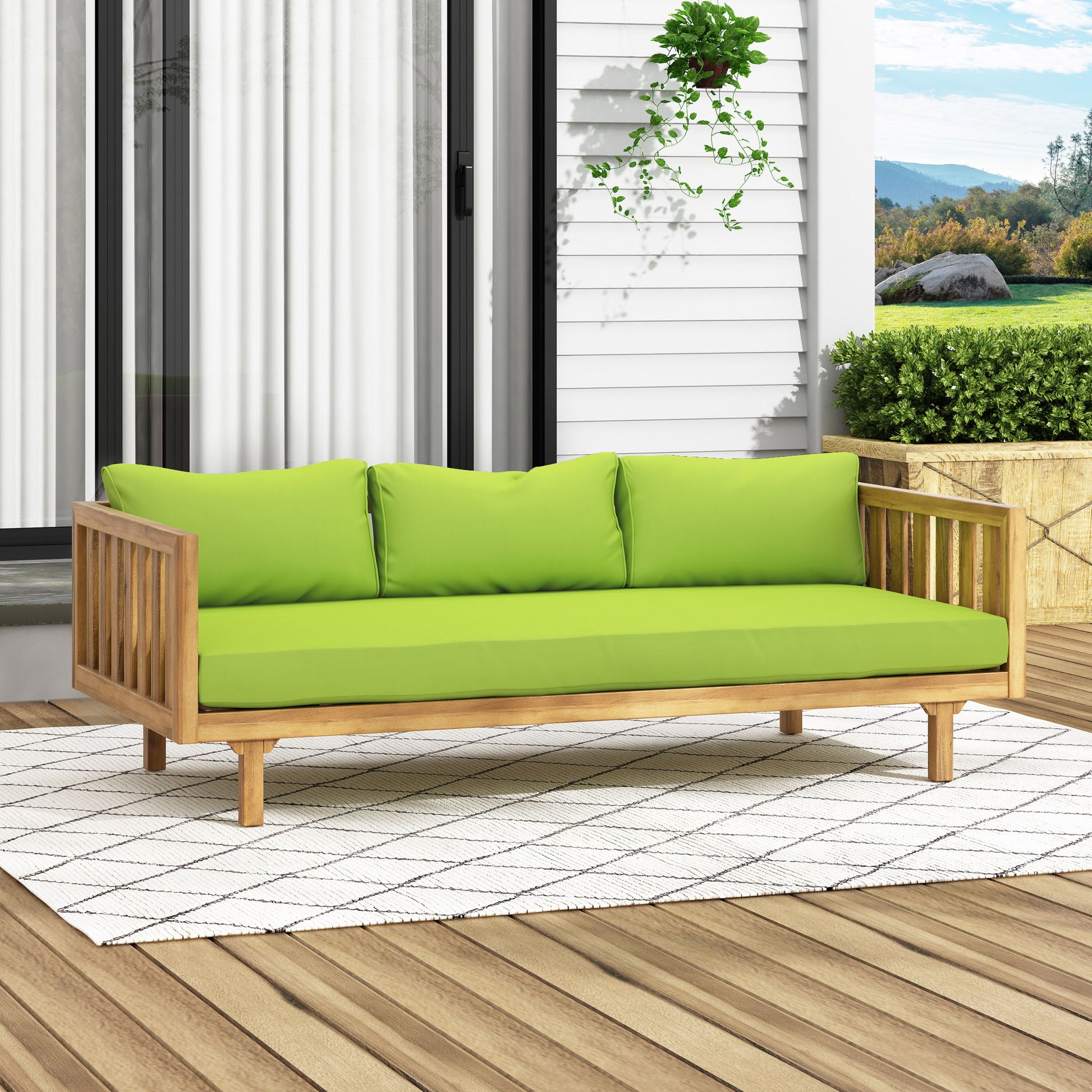 The Claremont - 3 Seater Daybed, crafted from teak and acacia wood with water-resistant green cushions and three pillows, rests on a white patterned rug on a wooden deck surrounded by plants, with a house and mountains in the background.