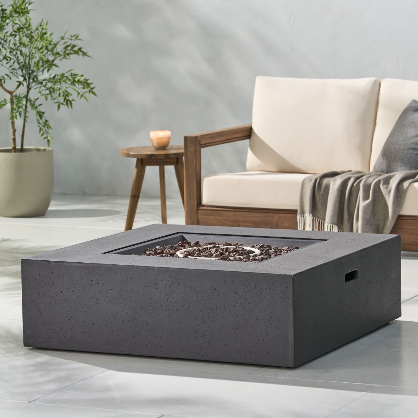 A modern outdoor patio with a beige cushioned sofa, wooden armrest, potted plant, gray blanket, and the Modern 50,000 Btu Square Fire Table With Propane Tank filled with dark rocks on a light stone floor.