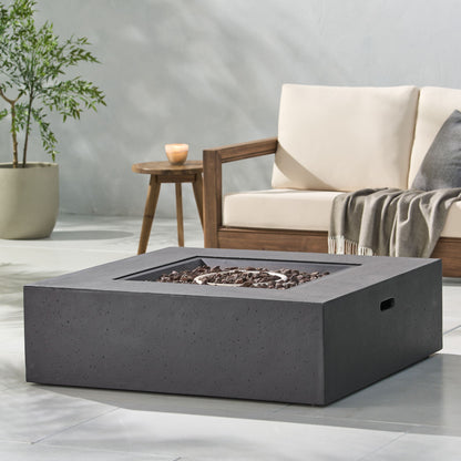 A modern outdoor patio with a beige cushioned sofa, wooden armrest, potted plant, gray blanket, and the Modern 50,000 Btu Square Fire Table With Propane Tank filled with dark rocks on a light stone floor.