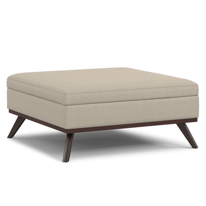 The Owen Multifunctional Square Coffee Table Storage Ottoman features light beige upholstery, a thick cushioned top, and dark wooden angled legs for a modern touch.