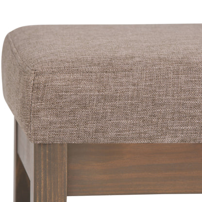 Close-up of the Milltown - Upholstered Ottoman Bench, highlighting its light brown textured fabric seat and wooden legs at the corner and side.