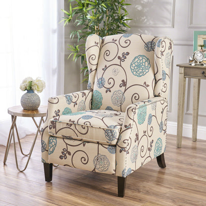 The Westeros Wing Back Fabric Upholstered Recliner, featuring blue and brown floral patterns, sits on a wooden floor beside a round side table with a vase of white flowers, while a potted plant and framed photo enhance the background.