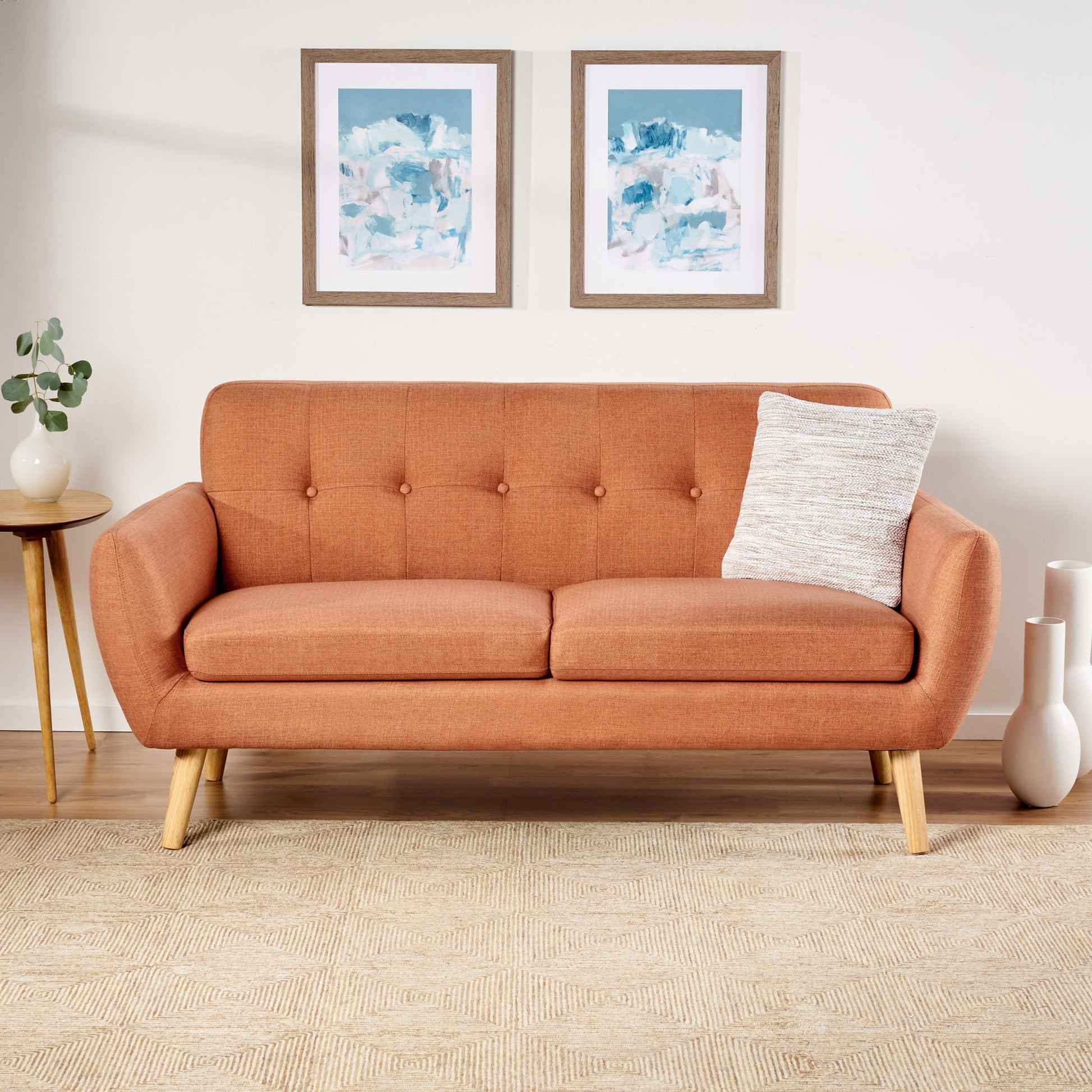A Modern Loveseat Sofa With Button Tufted Backrest featuring wooden legs and an orange finish sits by a white wall, paired with a gray textured pillow. Abstract art hangs above, while a side table and white vases rest on a light patterned rug.