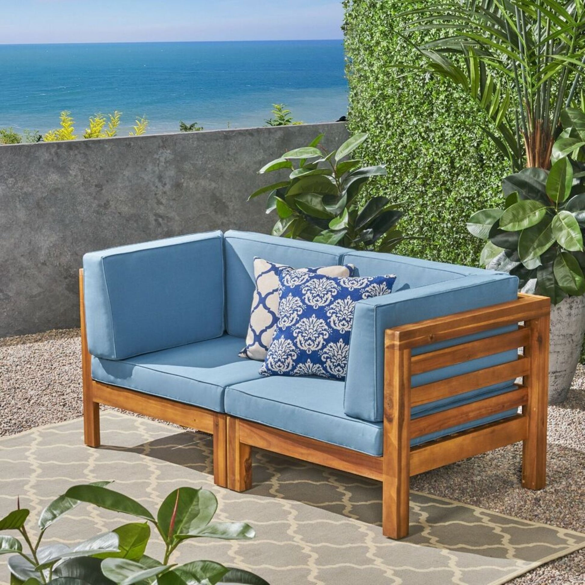 The Oana 2 Piece Loveseat Set with water-resistant cushions sits on a patio surrounded by potted plants, offering ocean and blue sky views.