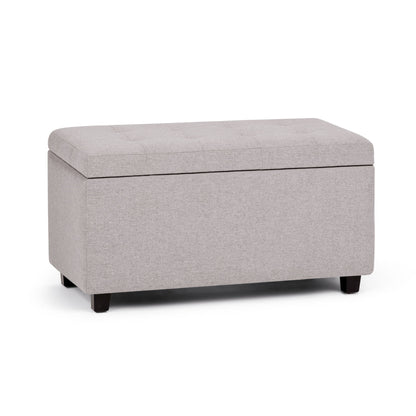 The Cosmopolitan Upholstered Storage Ottoman features a rectangular light gray design, cushioned tufted lid, and short dark wooden legs, shown against a white background.