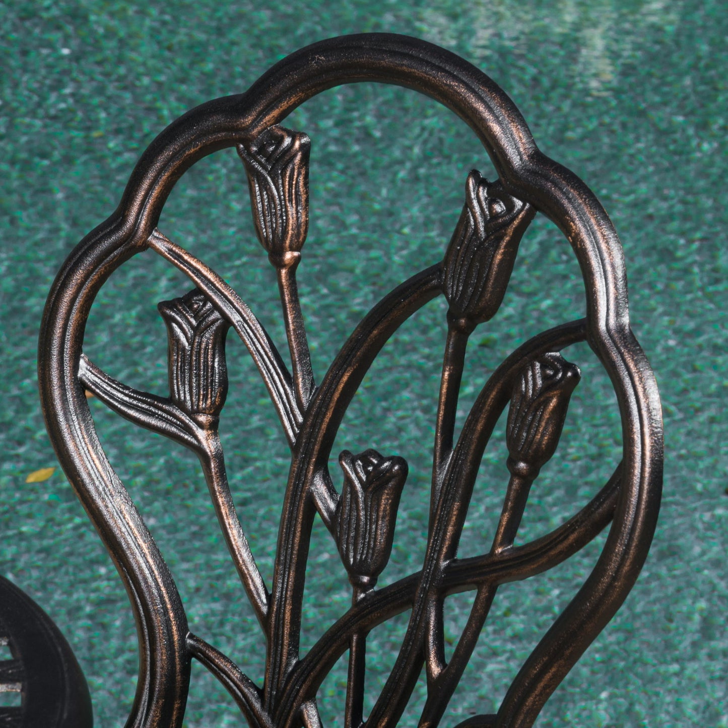 Close-up of the Nassau - Cast 3 Piece Bistro Set metal chair backrest, featuring a decorative tulip flower design, set against a textured green background.