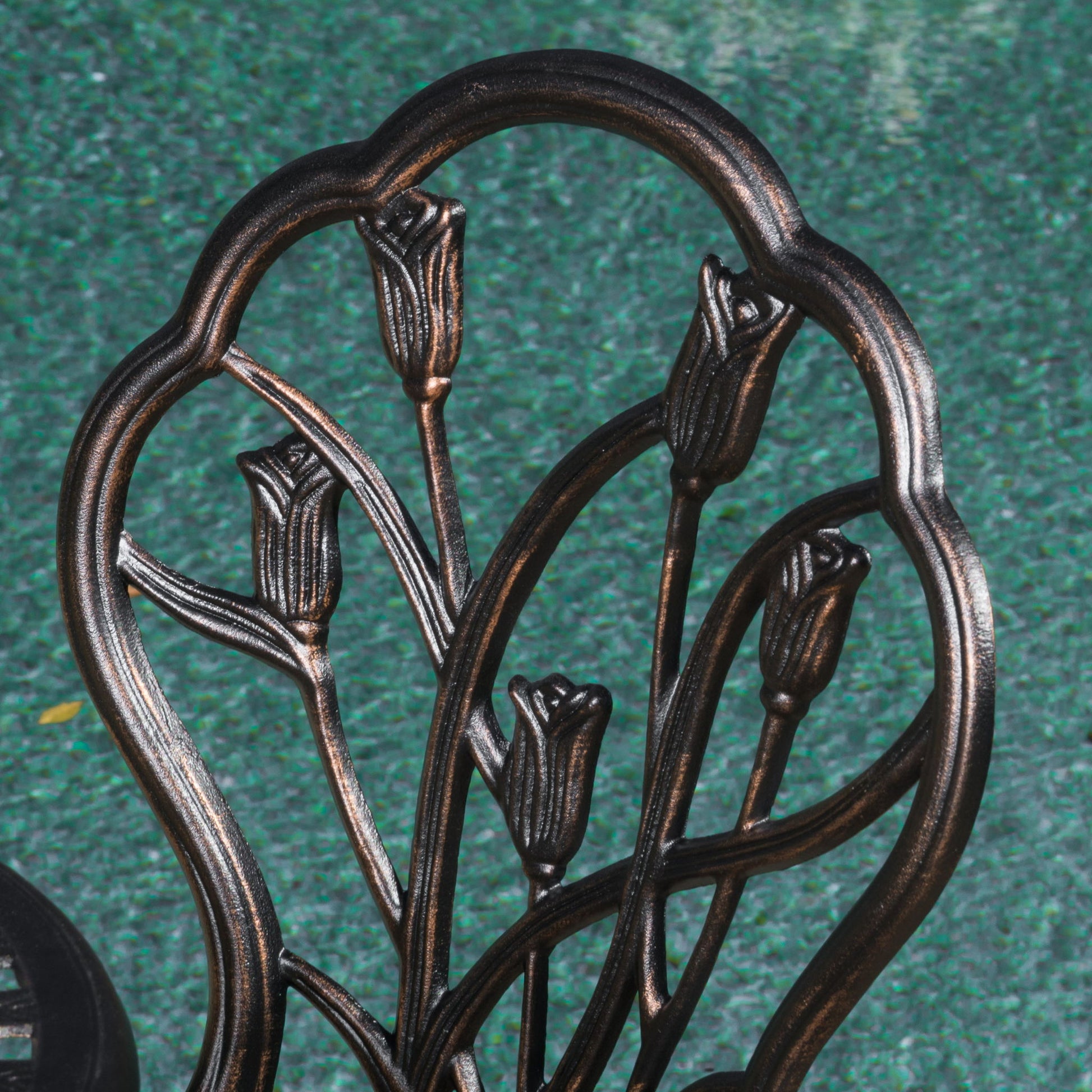 Close-up of the Nassau - Cast 3 Piece Bistro Set metal chair backrest, featuring a decorative tulip flower design, set against a textured green background.