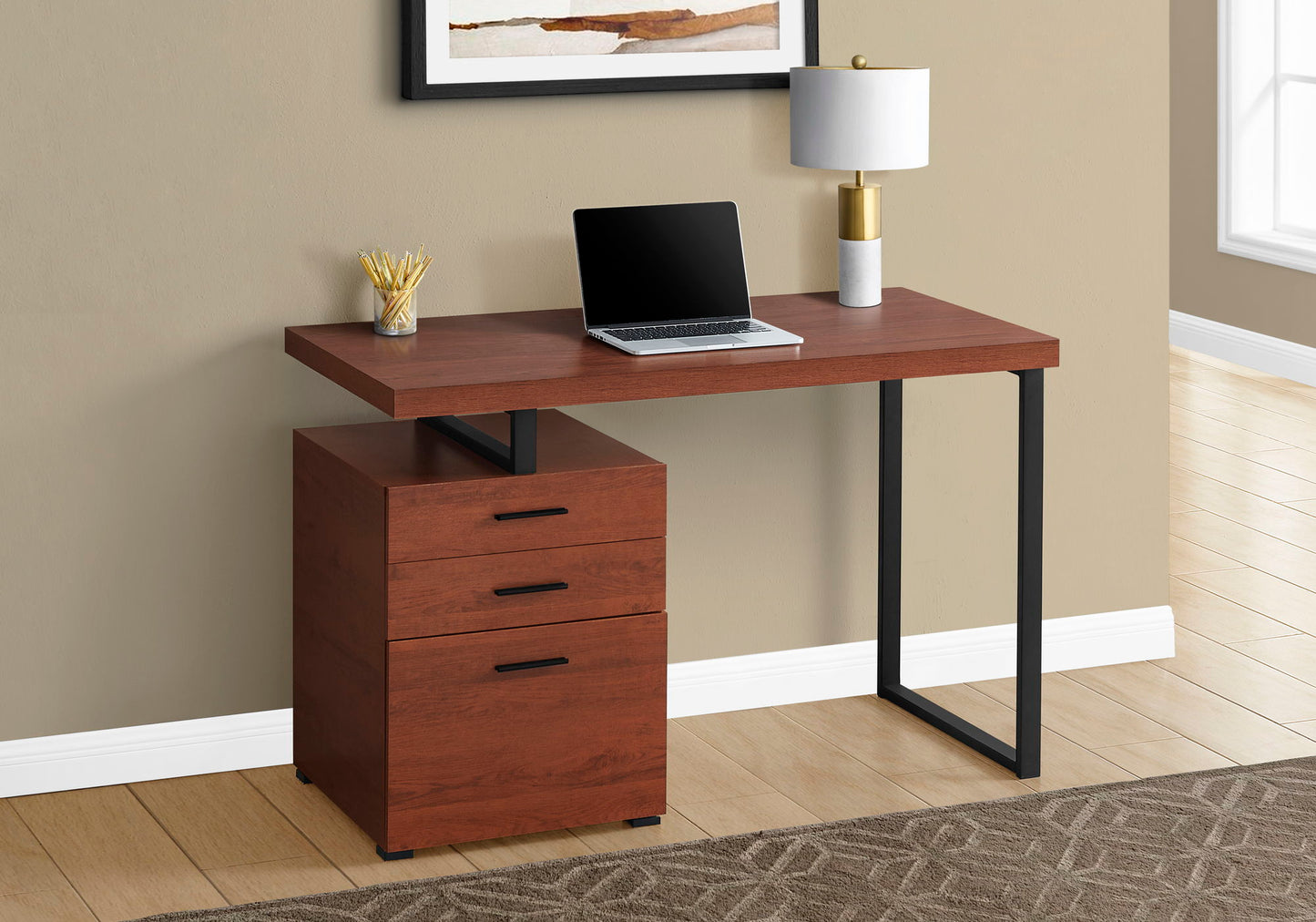 A Computer Desk For Home Office with storage drawers on the left, sleek black metal legs, and a wood finish holds a laptop, lamp, and pencil holder. It sits against a beige wall topped with framed artwork.