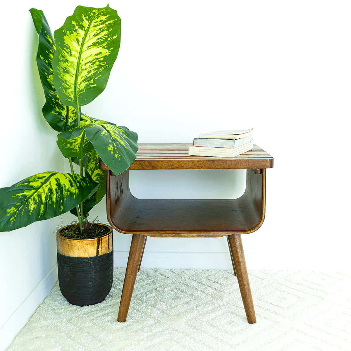 The Alice - Mid-Century Modern Table with books on top sits beside a tall green plant in a black and gold pot, all placed on a light patterned rug against a white wall.