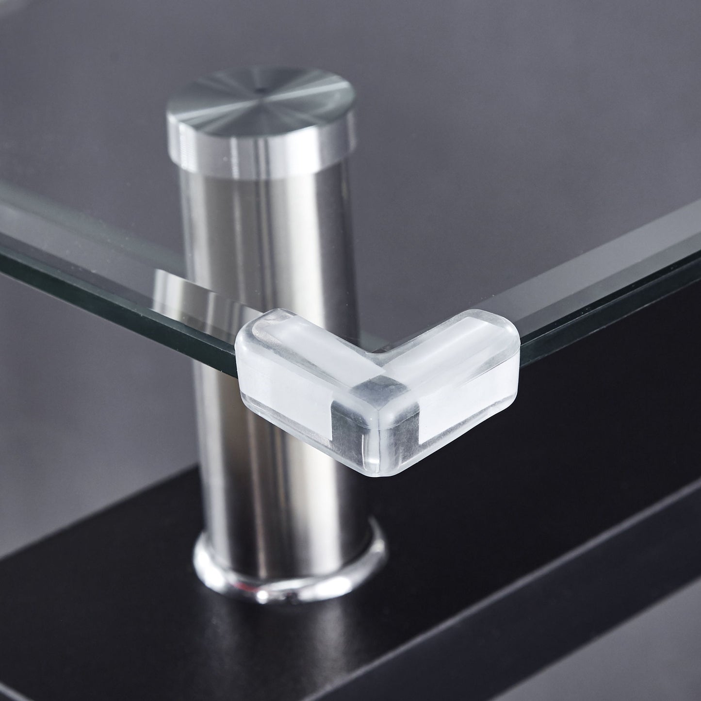 A Clear Glass Top Side Table with a modern design features a transparent corner protector on its edge and a metal support post behind the glass, set against a blurred, dark background.
