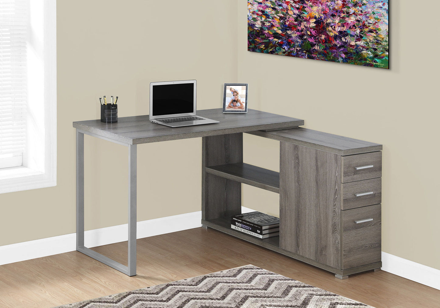 A contemporary gray L-shaped Computer Desk for the home office features storage drawers and space for a laptop. It sits in a bright room with beige walls, a colorful painting, and items such as a framed photo and cup of pens on top.