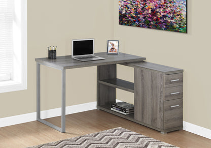 A contemporary gray L-shaped Computer Desk for the home office features storage drawers and space for a laptop. It sits in a bright room with beige walls, a colorful painting, and items such as a framed photo and cup of pens on top.