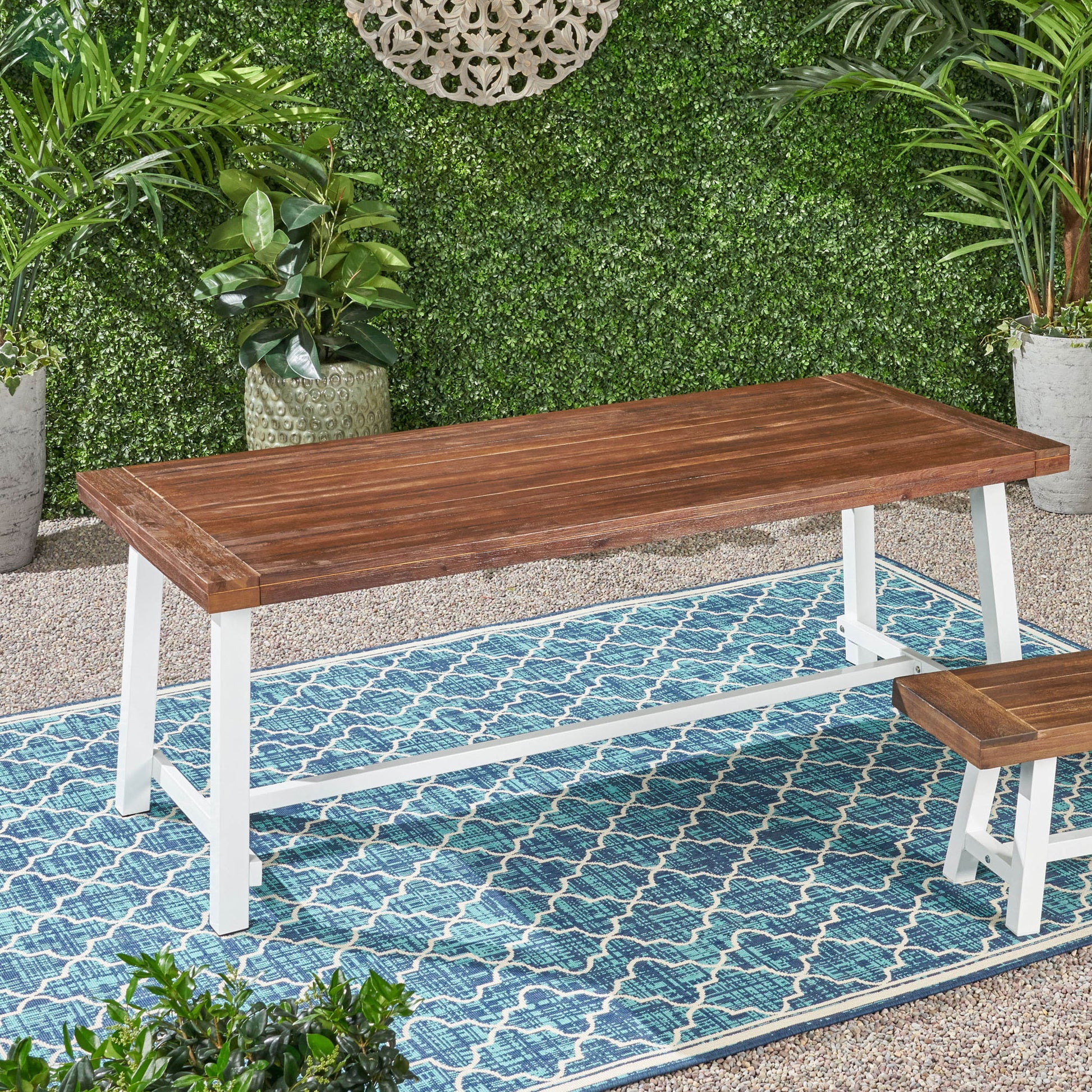 The Equilibrium Outdoor Dining Table, featuring a wooden frame and slat top with white legs, is set on a blue patterned rug among green plants and a leafy wall, creating a cozy garden vibe.