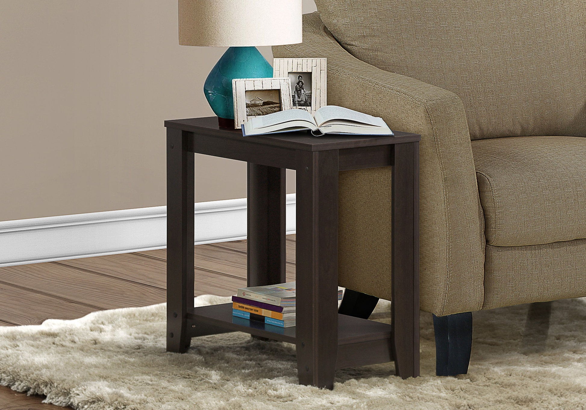 An Accent Table, Side Transitional with an elegant design stands by a beige sofa, topped with a blue lamp, two framed photos, and an open book. More books are stored on its lower shelf, all set on a light-colored rug.