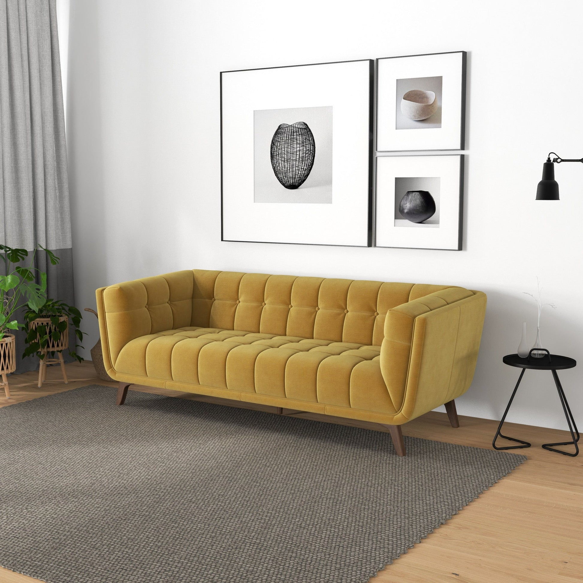 A modern living room featuring the Addison Mid-Century Modern Tufted Sofa in mustard yellow on a gray rug, a black side table with a lamp, minimalist wall art prints, gray curtains, and a green plant in the corner.