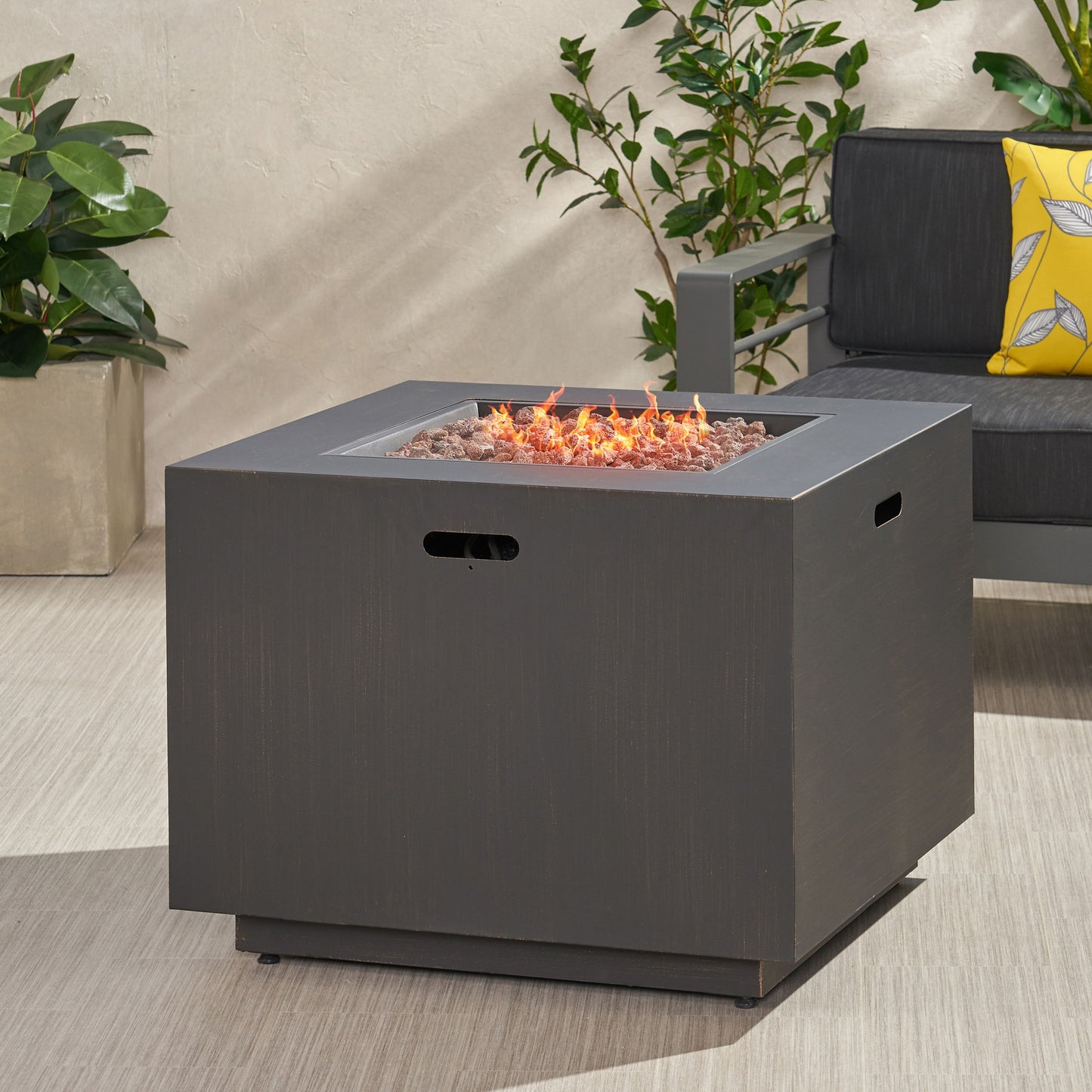 The Outdoor Patio Gas Burning Fire Pit Table 50,000 Btu, Square, features visible flames and is placed on a patio beside gray outdoor furniture with a yellow pillow and green potted plants in the background.