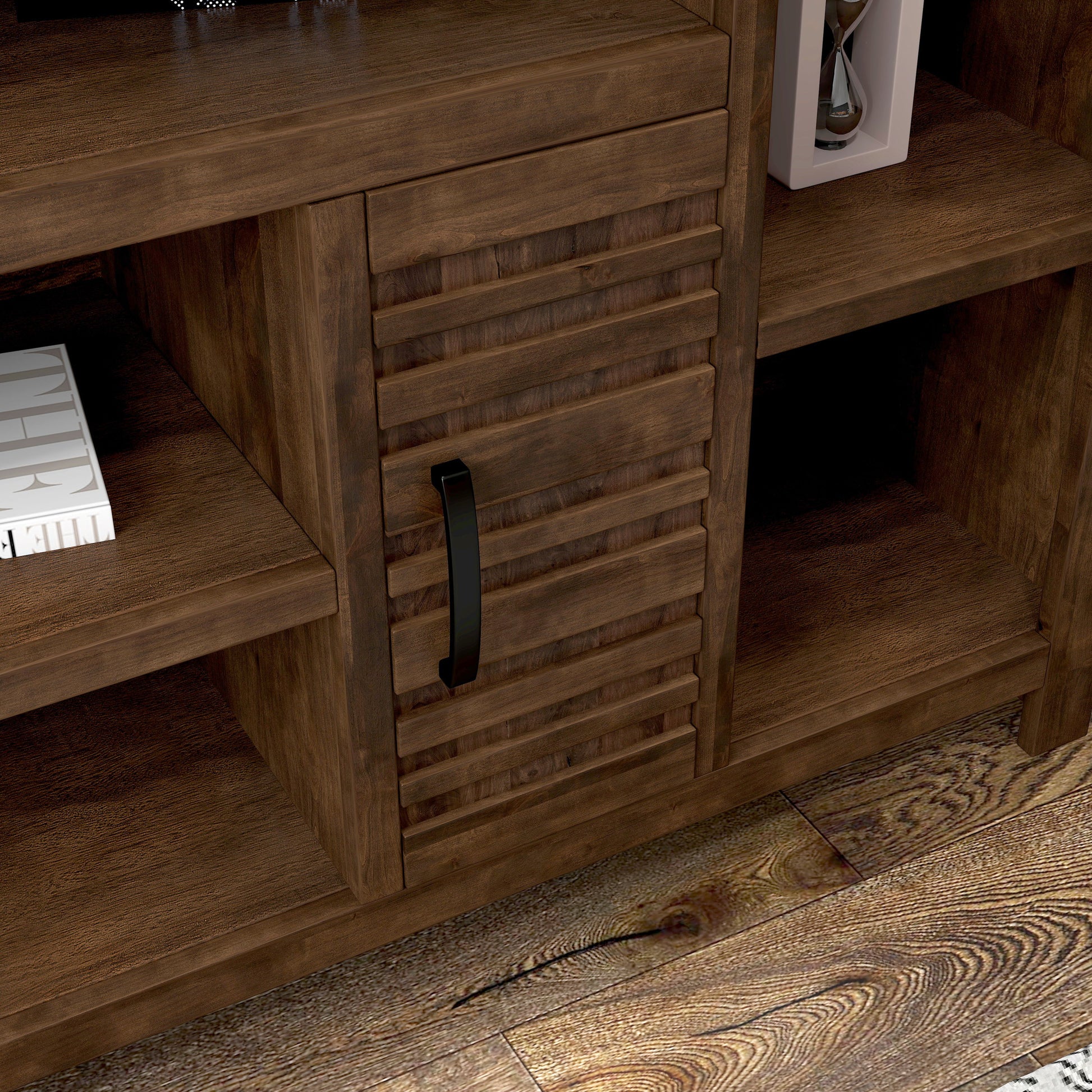 A close-up of the Sausalito - TV Stand Console For TVs features a slatted wooden door with a black handle, open shelving displaying books and an hourglass, all set on a wooden floor.
