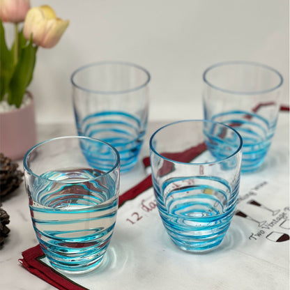 A set of four Leading Ware Swirl Acrylic Drinking Glasses is arranged on a white surface with red accents, accompanied by a pink pot of yellow and pink tulips and some pinecones in the background.