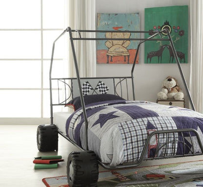 A kids' bedroom featuring the Xander Twin Go Kart Bed in gunmetal, star-patterned bedding, a large window, wall art, and a stuffed animal on a chair. Books lie scattered on the floor.