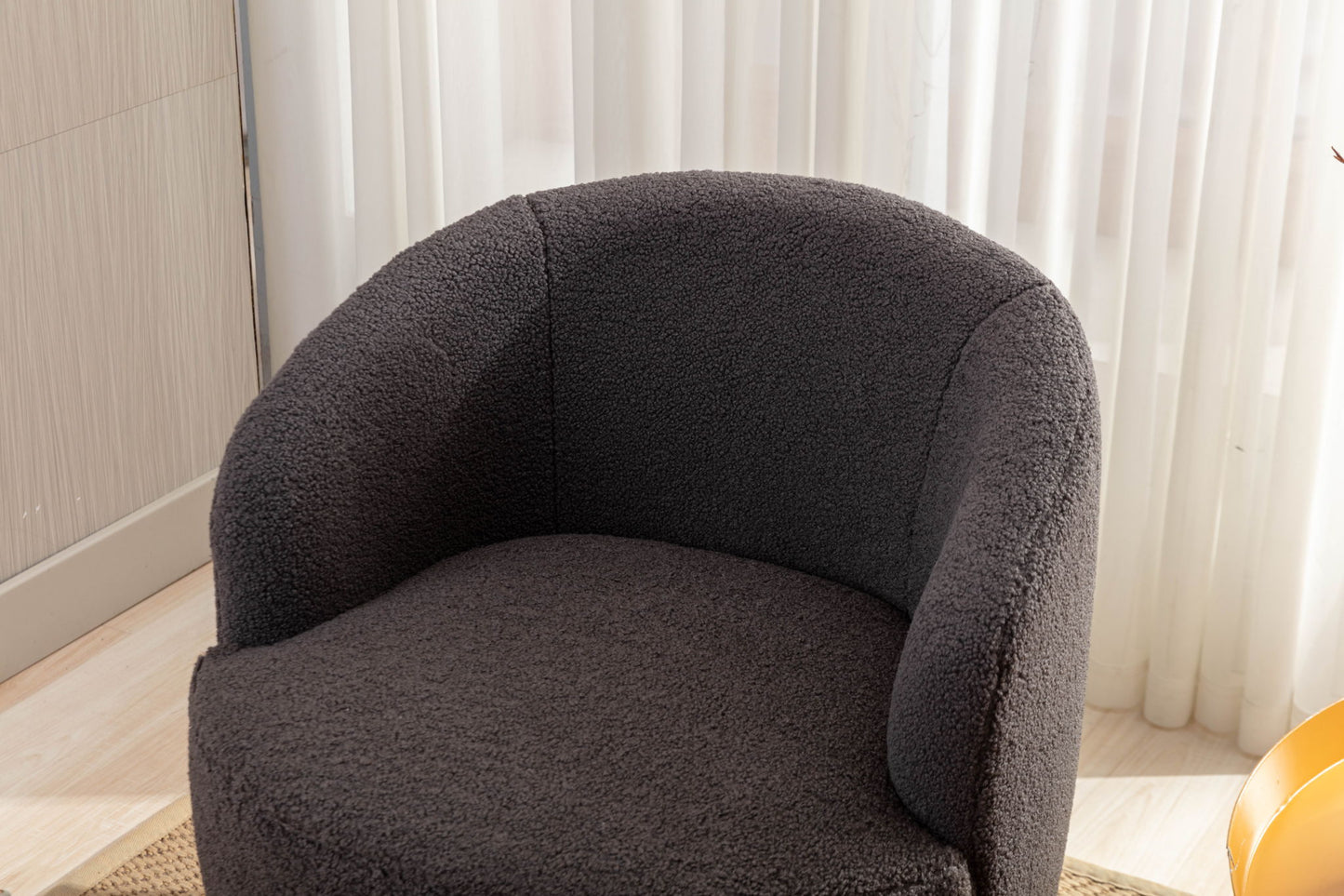The Teddy Fabric Swivel Accent Armchair Barrel Chair with a powder-coated metal ring, in modern dark gray textured fabric, sits on a light wood floor before white sheer curtains.