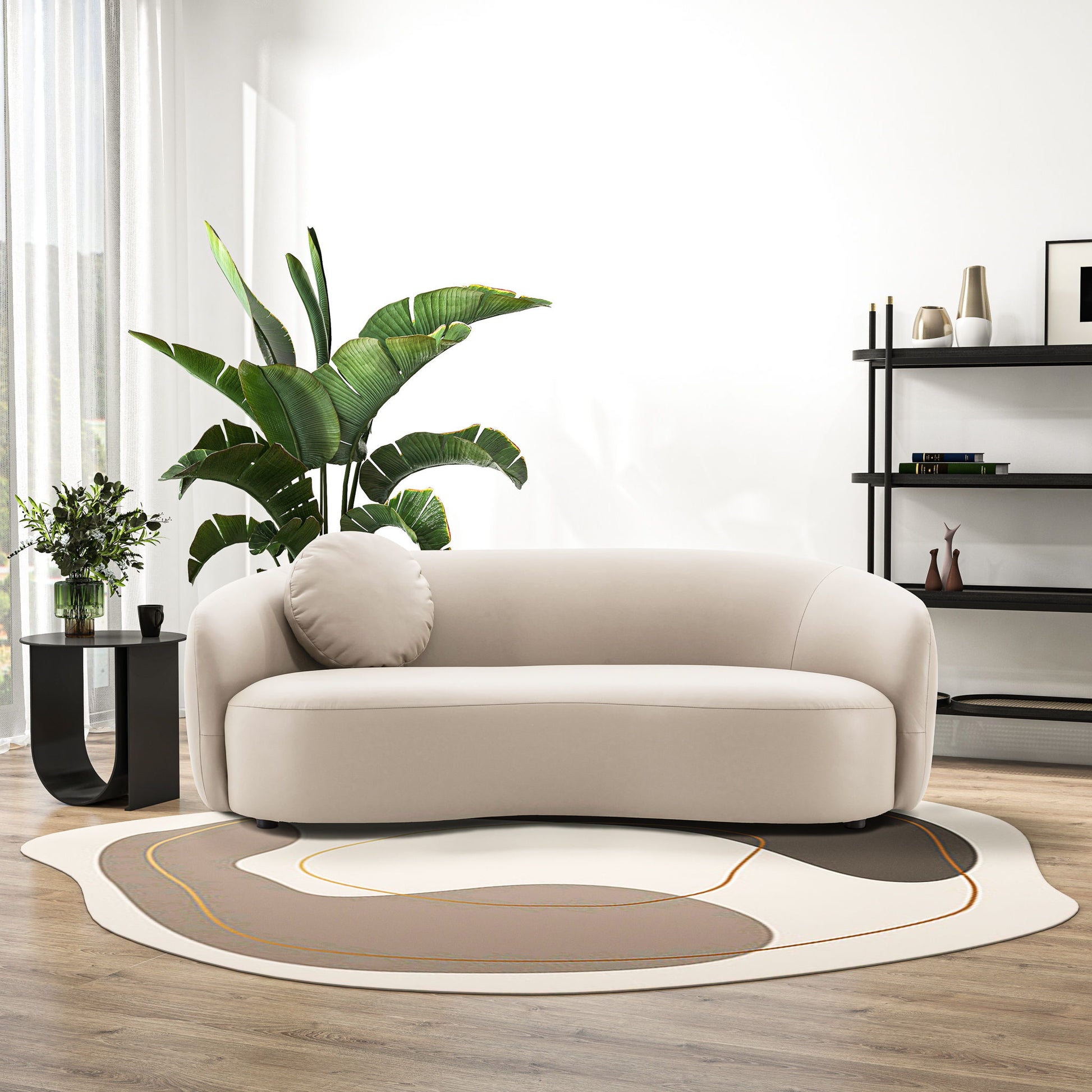 A modern living room features the Porter - Comfortable Sofa in beige, round pillow, abstract rug, black side table with a plant, tall green plant, and a black shelving unit on wooden floors. Soft light filters through sheer curtains against a white wall.