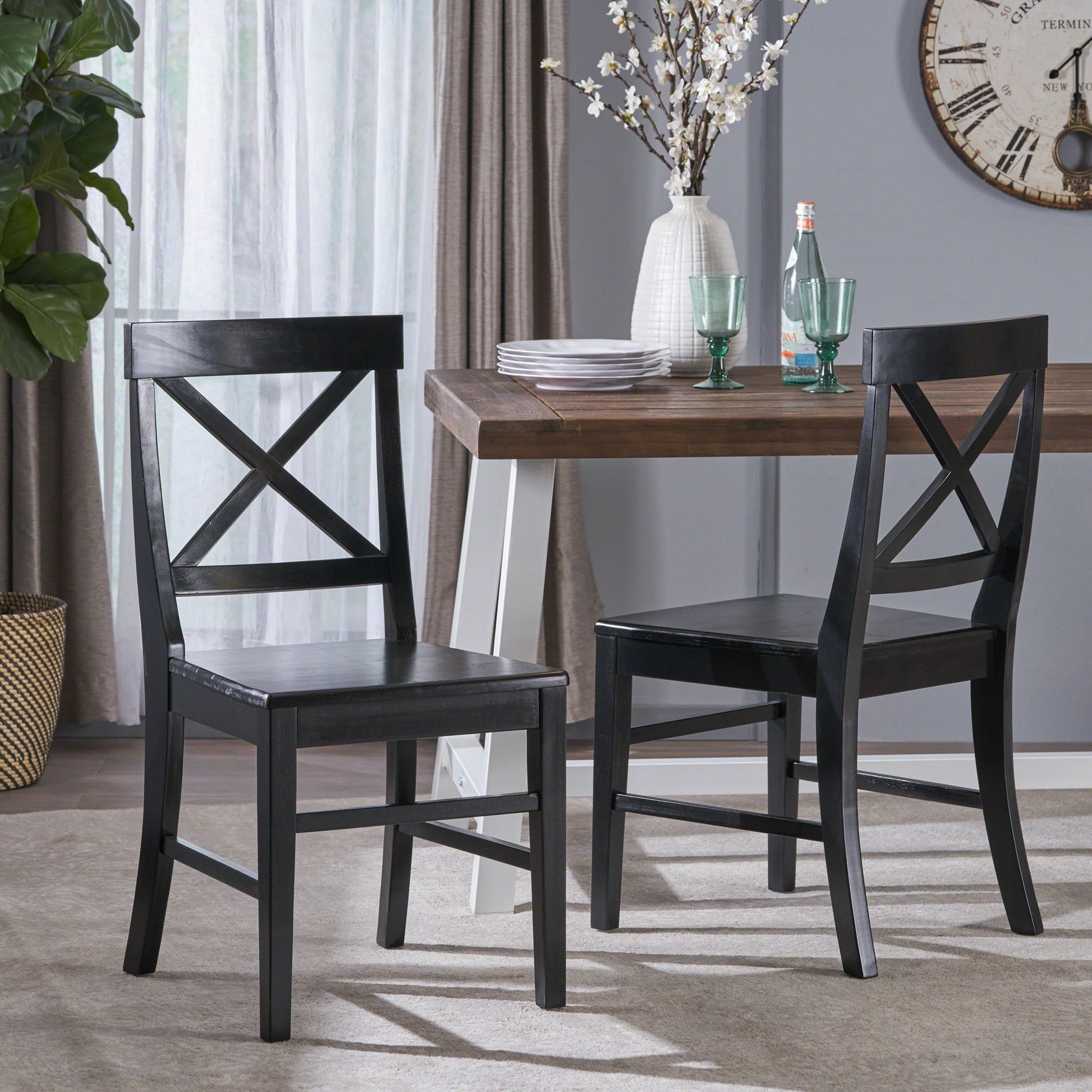 Two Plethora Dining Chairs with X-shaped backs (set of 2) sit by a wooden dining table topped with plates, glasses, a white vase with flowers, and a bottle. A large wall clock and curtains are visible in the background.
