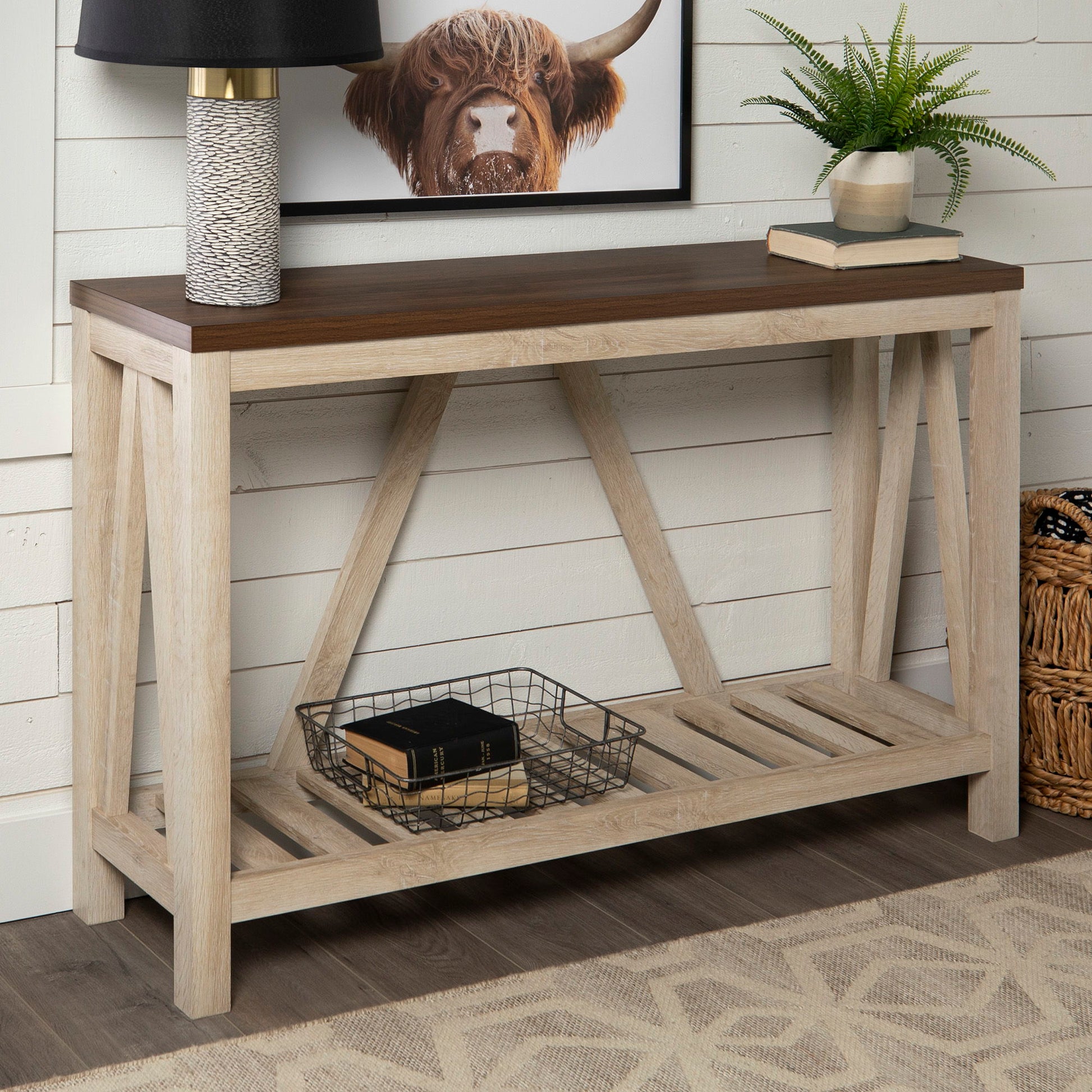 The Farmhouse A Frame Entry Table With Lower Shelf featuring a brown top and light base sits by a white shiplap wall, styled with a lamp, plant, highland cow art above, and a wire basket with books on the lower shelf.