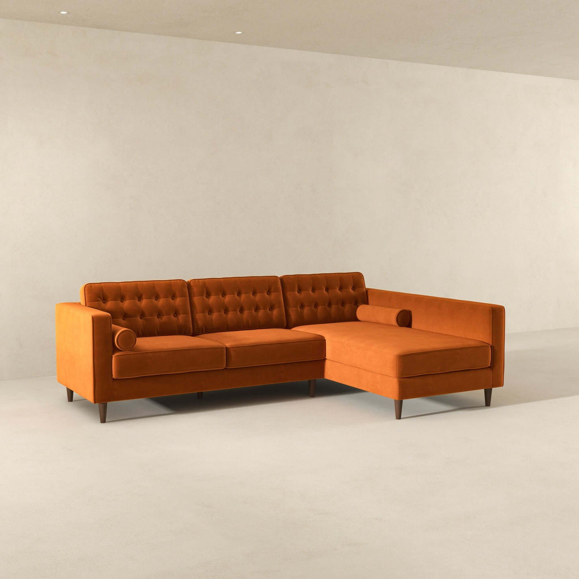 The Christian Mid-Century Modern Design Sectional Sofa, featuring tufted cushions and round bolster pillows in orange, is showcased in a minimalist room with light-colored walls and floors.