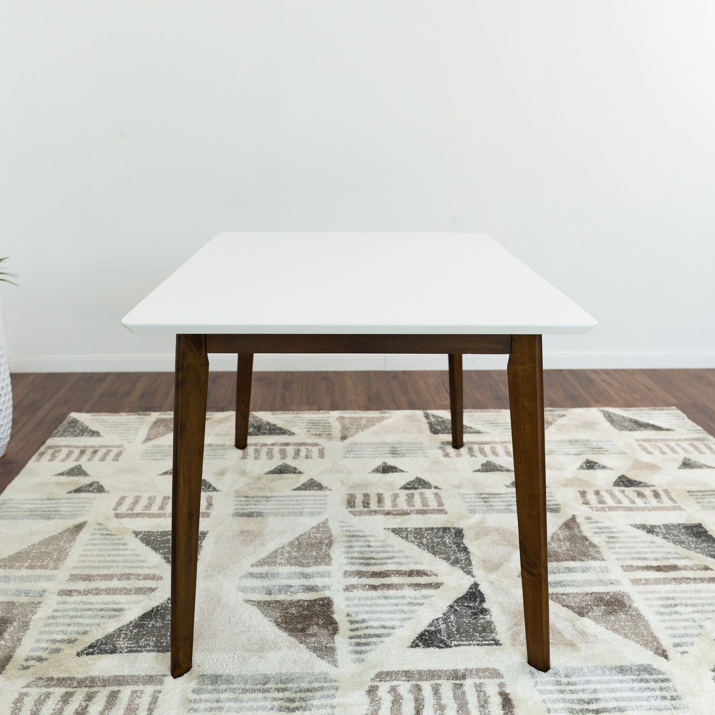 The Carlos Dining Table, featuring a white square top and wooden legs, is placed on a patterned neutral rug in a room with wooden floors and plain white walls.