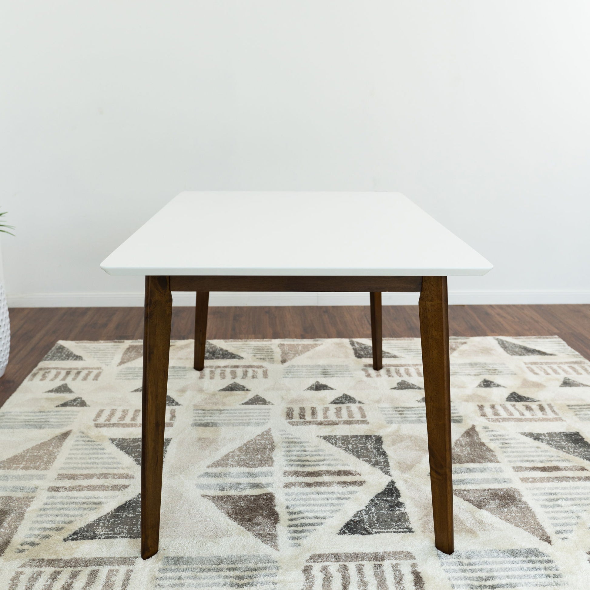 The Carlos Dining Table, featuring a white square top and wooden legs, is placed on a patterned neutral rug in a room with wooden floors and plain white walls.