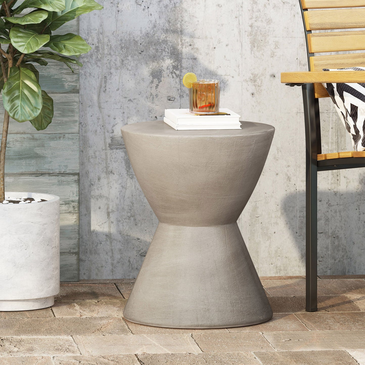 The Outdoor Hourglass Shaped Side Table in light gray holds a drink on a brick patio, next to a potted plant and a wooden chair with striped cushion, all set against a textured wall.