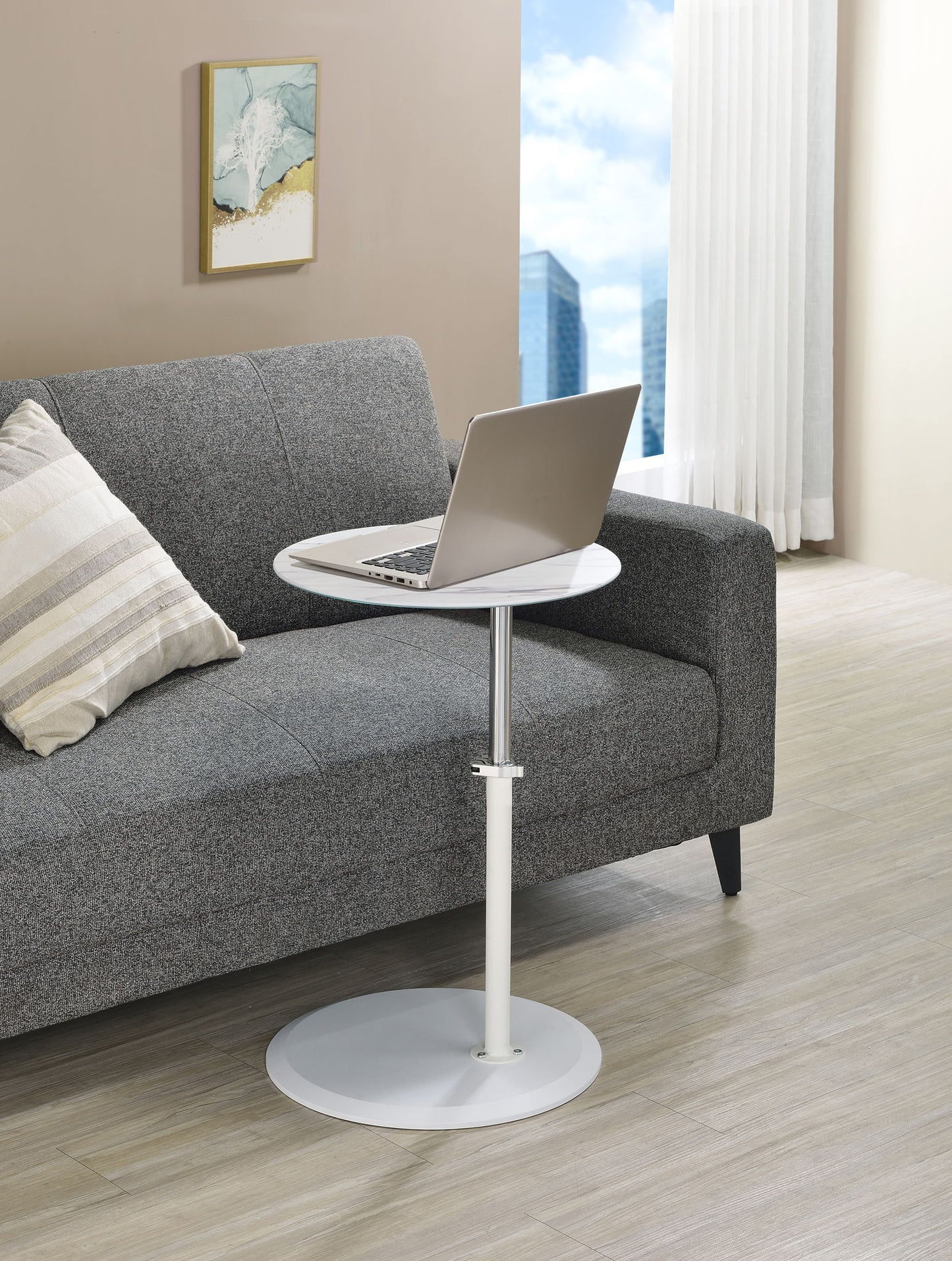 A modern living room with a gray sofa, striped pillow, laptop on the Orbit 15.5" End Table with height adjustable marble textured top, and a window displaying city buildings and blue sky.