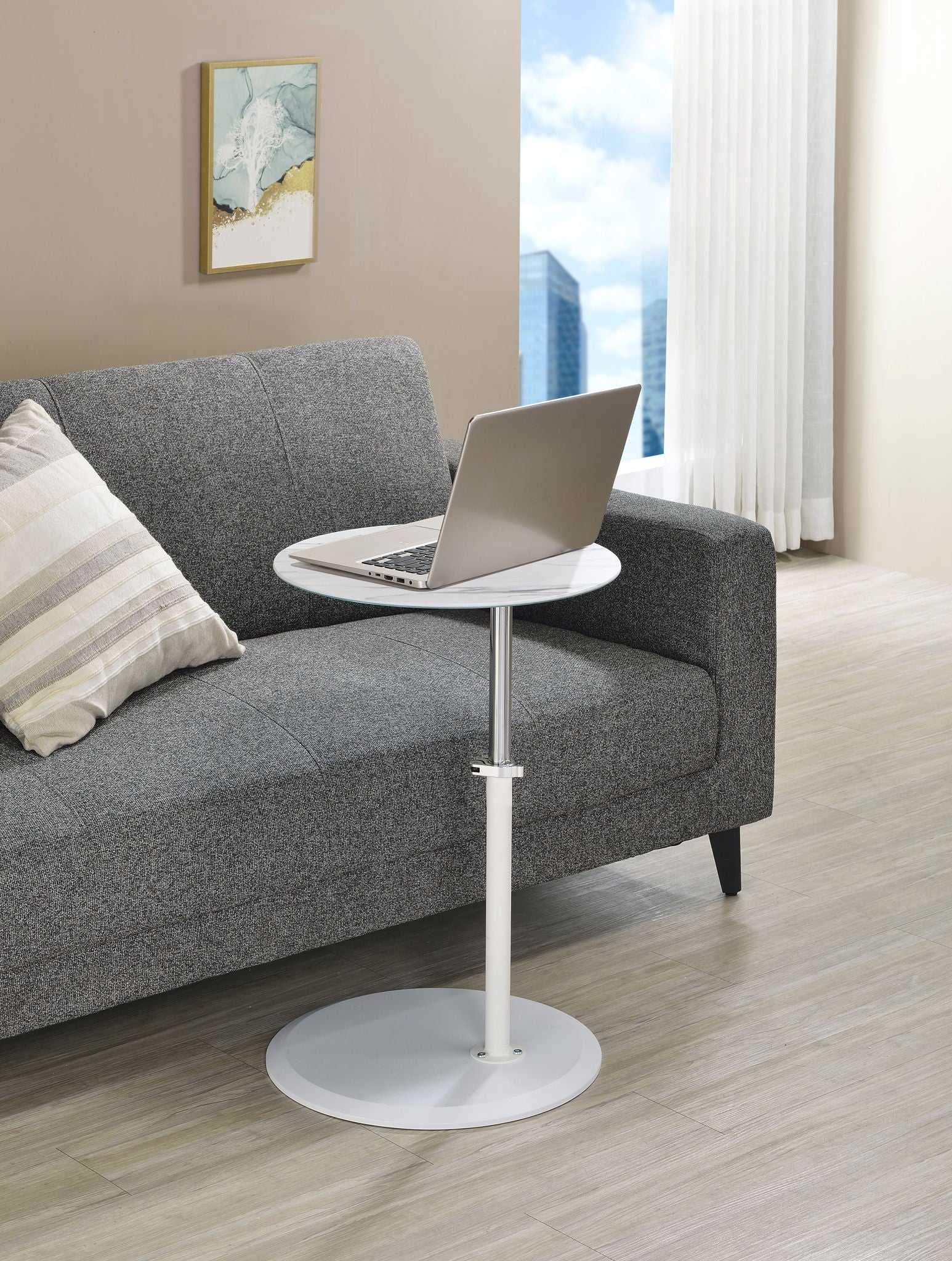 A modern living room with a gray sofa, striped pillow, laptop on the Orbit 15.5" End Table with height adjustable marble textured top, and a window displaying city buildings and blue sky.