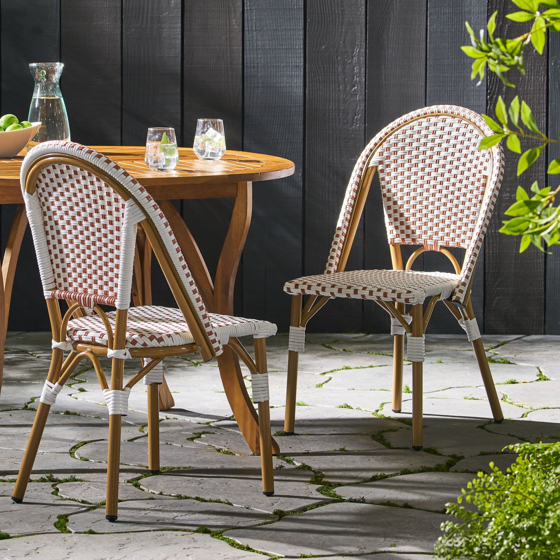 Two Outdoor Rattan and Aluminum French Bistro Chairs and a round table with glasses and a pitcher are arranged on a stone patio surrounded by greenery in gentle sunlight.