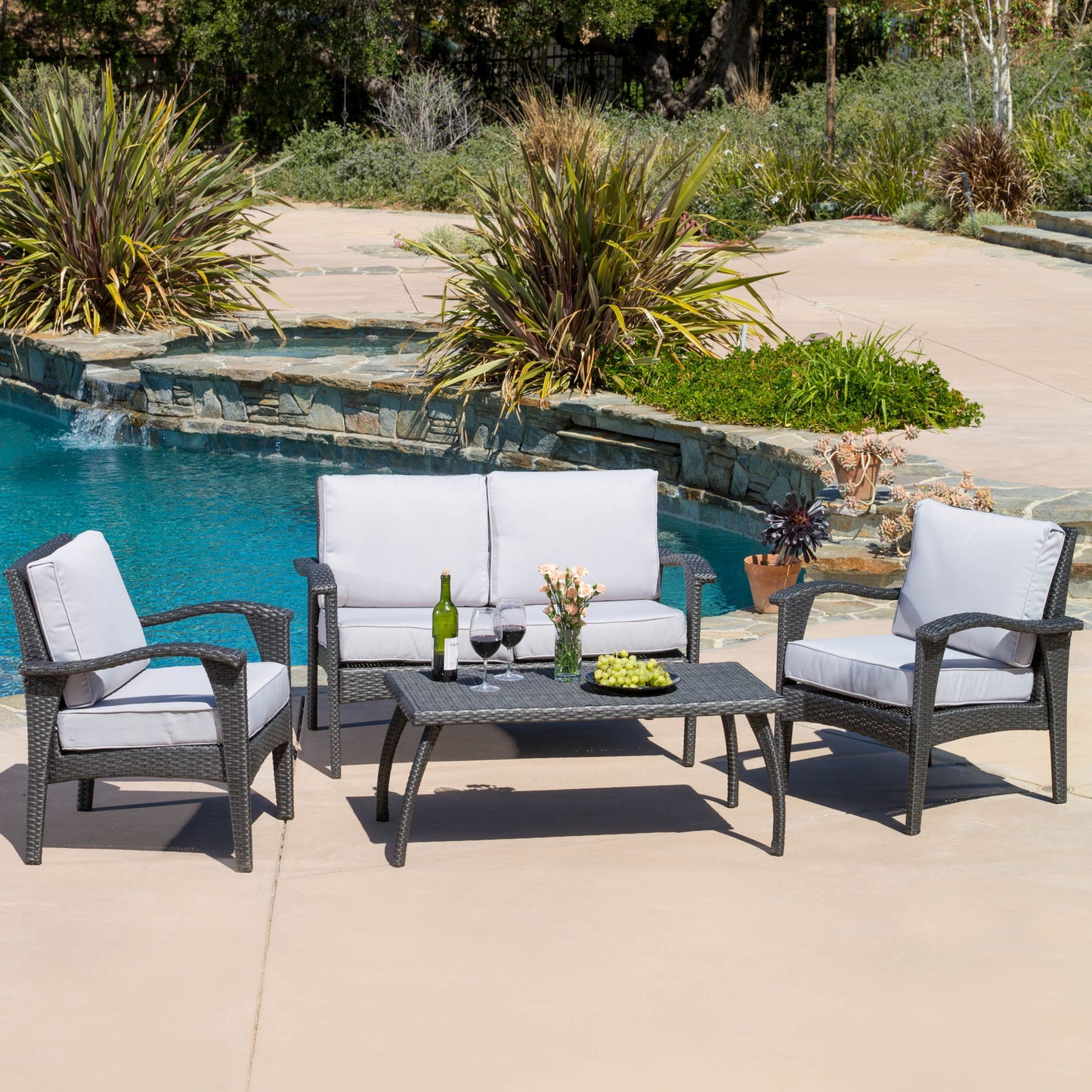 The Honolulu - Wicker Set Outdoor Seating, featuring gray-cushioned loveseat, two armchairs, and coffee table with wine, glasses, flowers, and snacks, is arranged by a pool surrounded by plants.
