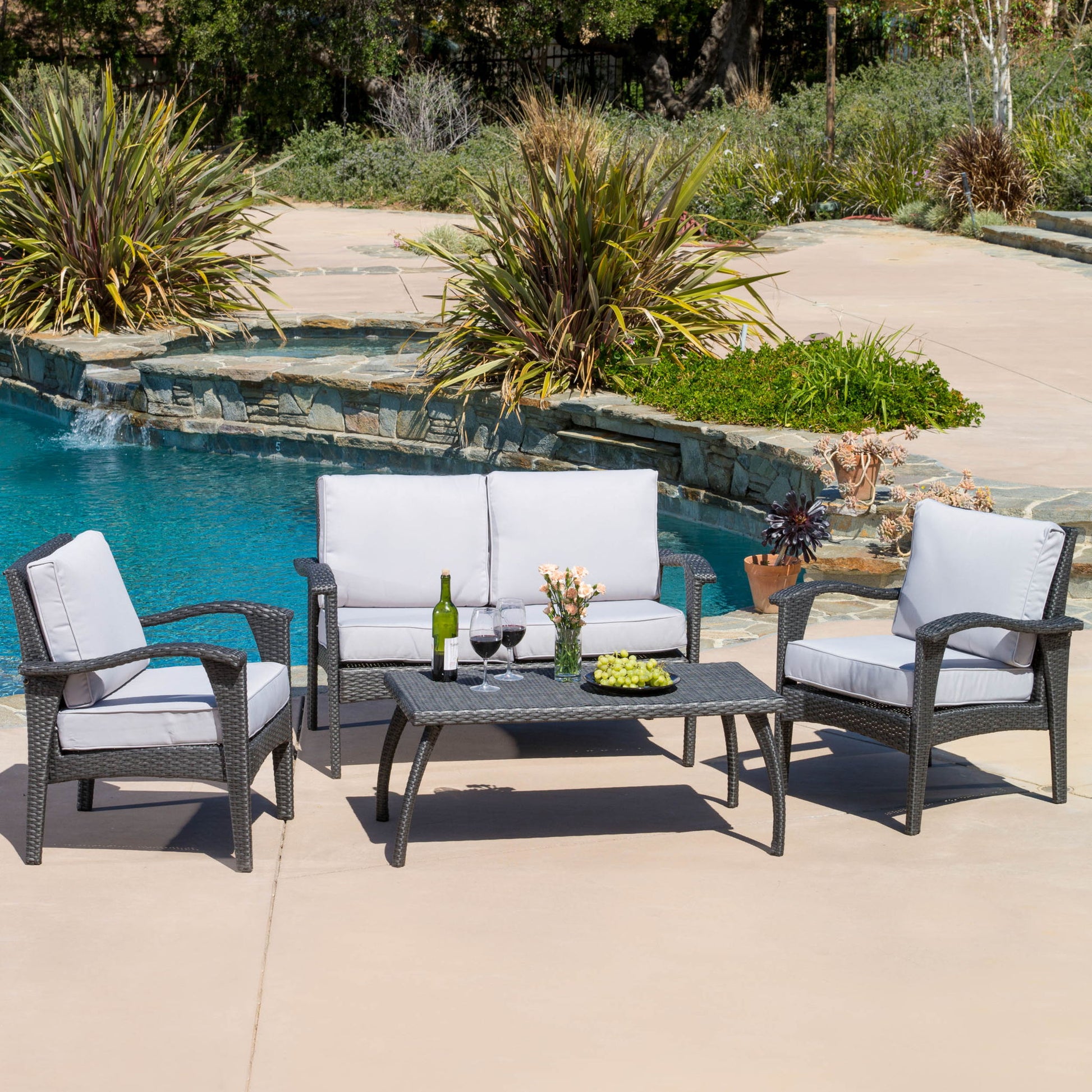 The Honolulu - Wicker Set Outdoor Seating, featuring gray-cushioned loveseat, two armchairs, and coffee table with wine, glasses, flowers, and snacks, is arranged by a pool surrounded by plants.